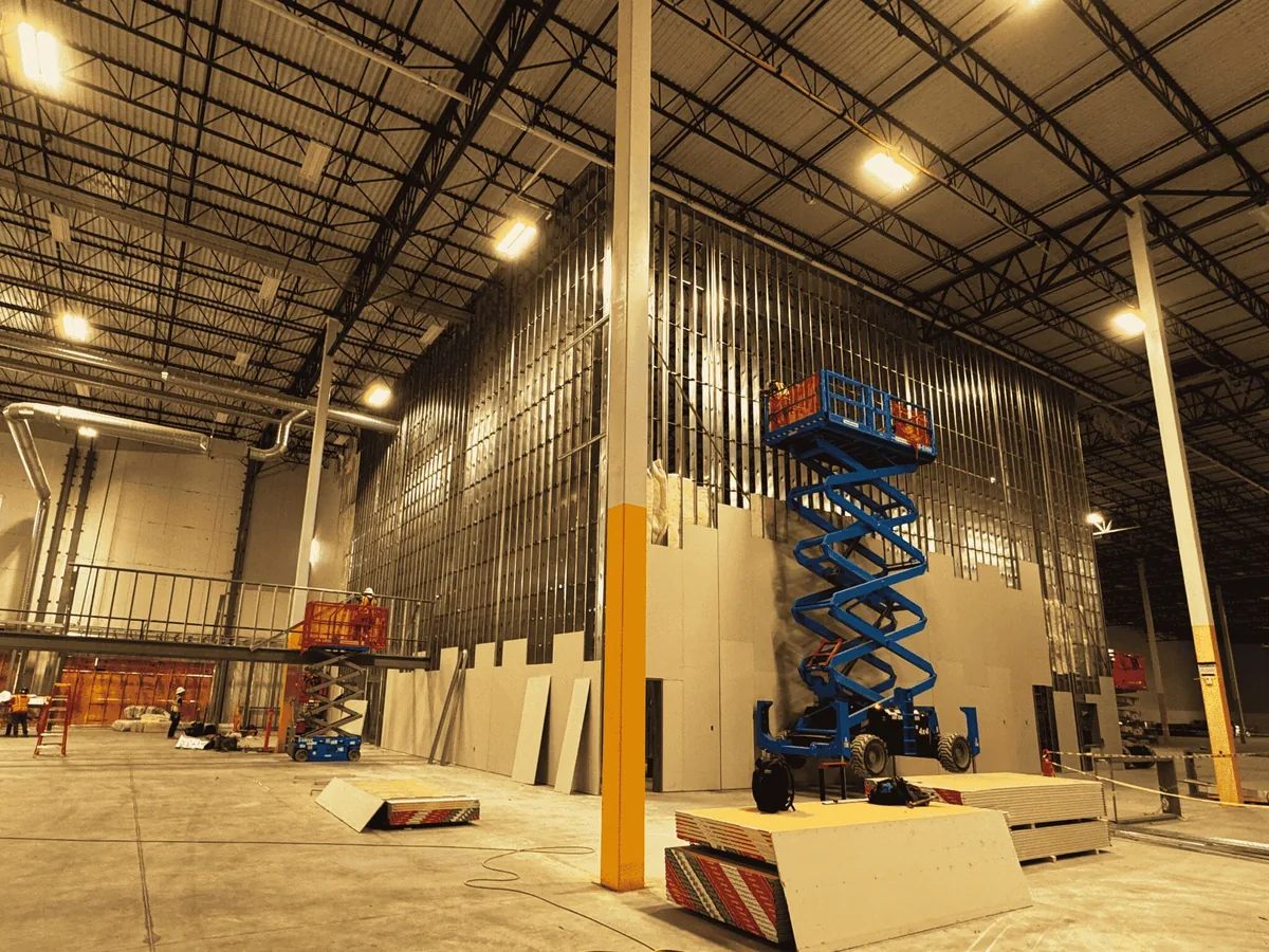 Tall metal stud wall being framed and drywalled inside a large warehouse with scissor lifts, commercial upfits