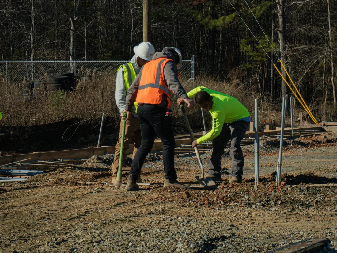 Three workers digging beside foundation forms and conduit on a cleared site, ground up construction