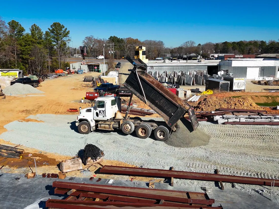 Dump truck spreading stone on a cleared commercial site with steel beams staged in the foreground, ground up construction