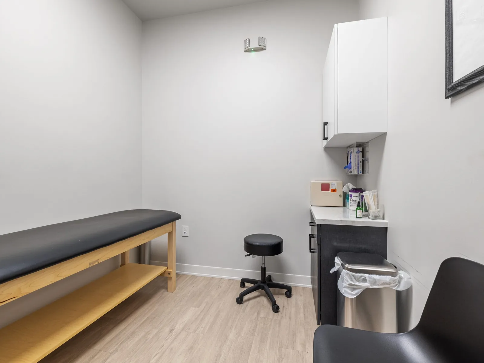 Small finished clinic exam room with a treatment table, a stool and a wall cabinet over a counter, medical office build outs