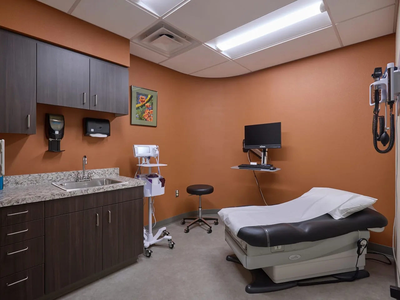 Finished clinic exam room with wood cabinets, a sink, an exam table and a wall mounted monitor, medical office build outs