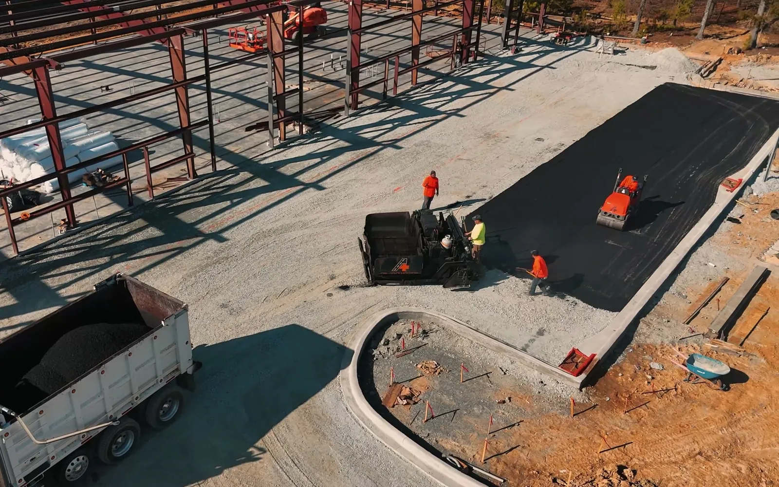 Aerial view of red steel building frames beside a fresh asphalt lot and a loaded dump truck, metal building construction