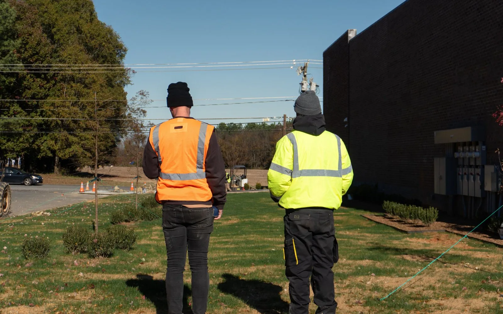 Two builders in safety vests reviewing a site beside a dark brick commercial building, preconstruction