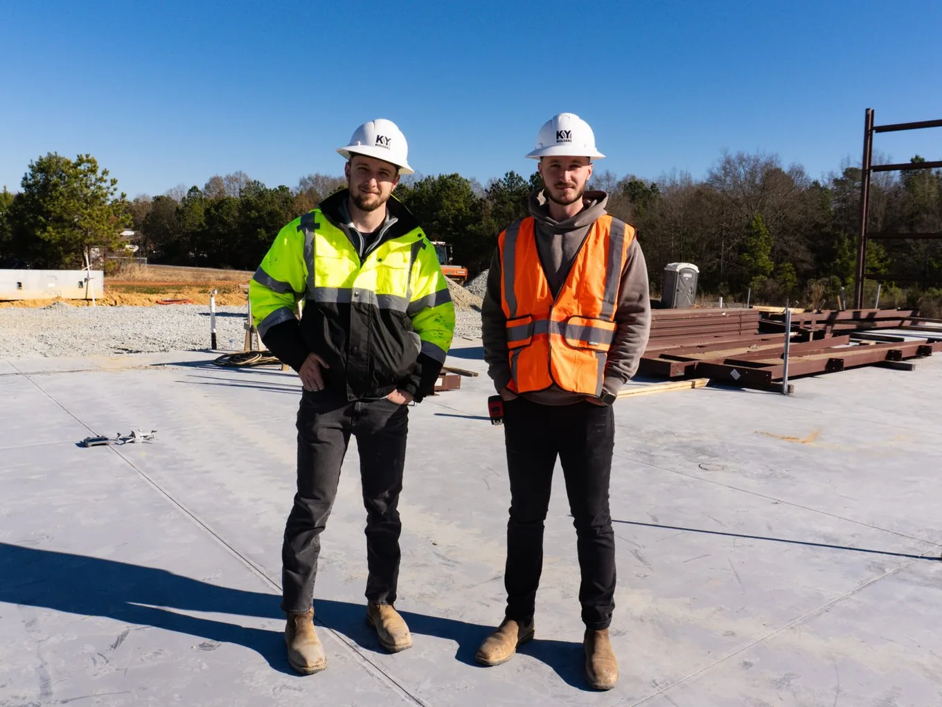 Two builders in hard hats and safety vests standing on a new building pad with stacked steel, commercial general contractor team