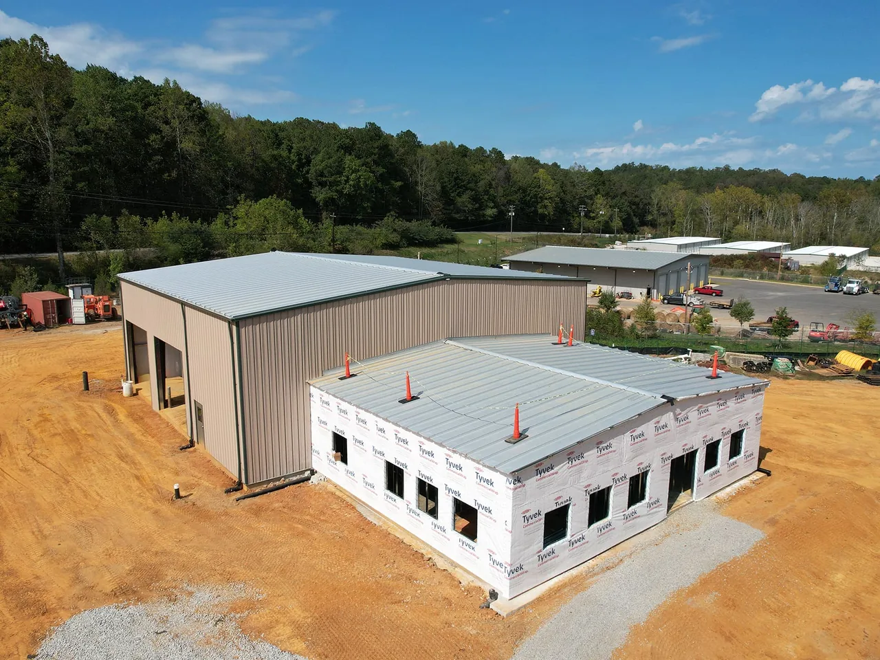 New tan metal warehouse with open door bays and a wrapped office wing going up on a graded red clay site, warehouse construction