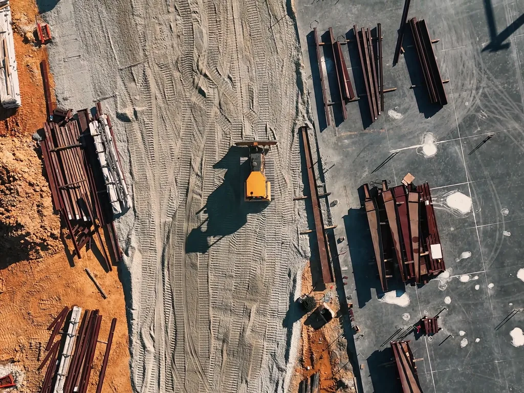 Overhead view of steel members staged on red clay beside a new slab, warehouse construction